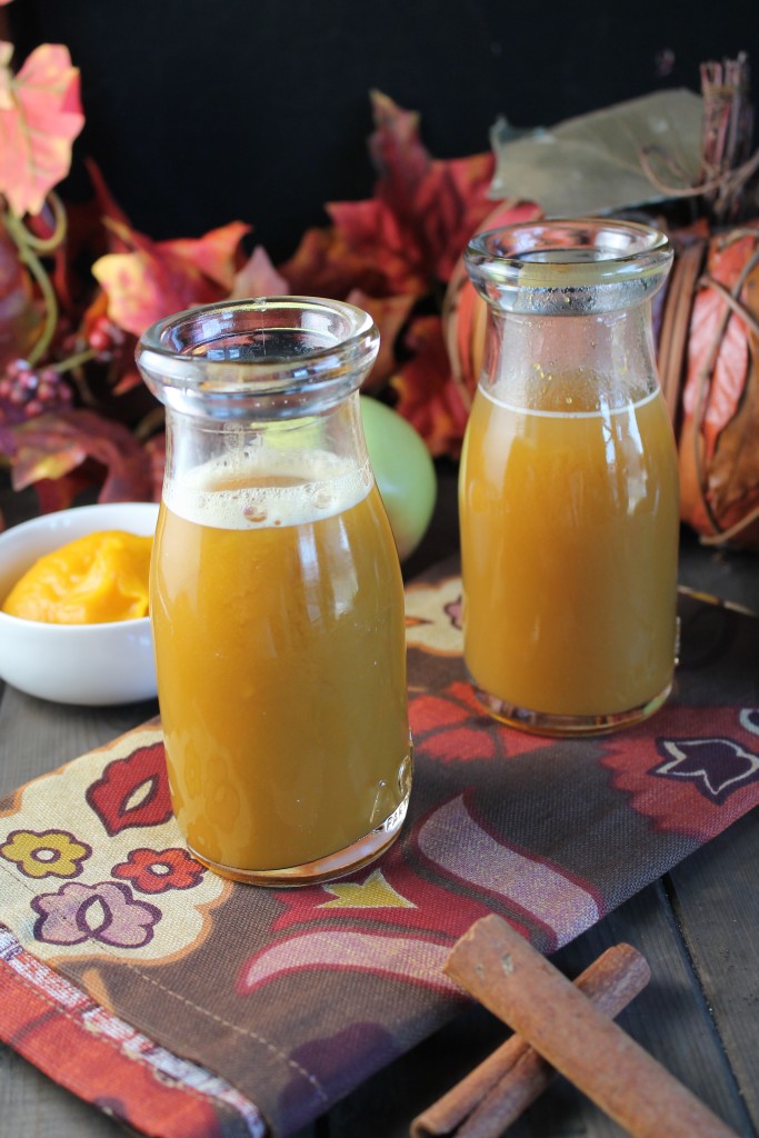 two clear glasses filled with pumpkin apple cider with fall themed cloth, dark wood surface, fresh apples, fresh pumpkin puree, and fall leaves
