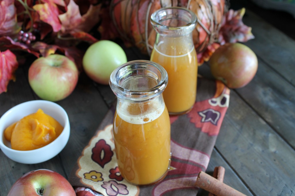 two vintage milk glasses filled with an orange hued apple cider drink on a dark wooden surface with fresh apples and fall leaves