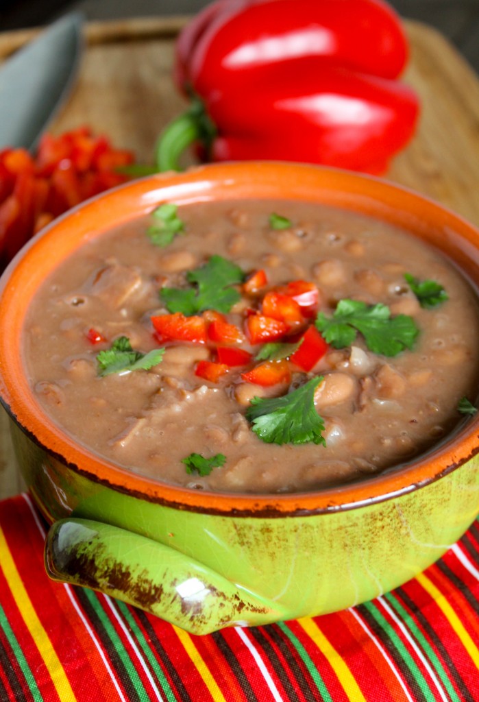 a green ceramic handled bowl filled with creamy cooked pinto beans with chopped red pepper and cilantro on top