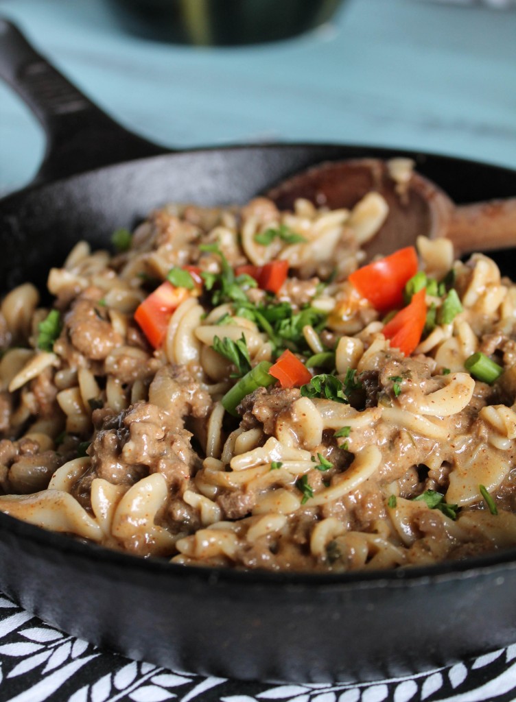 a cast iron skillet filled with a cheesy hamburger noodle dish garnished with fresh red pepper and parsley