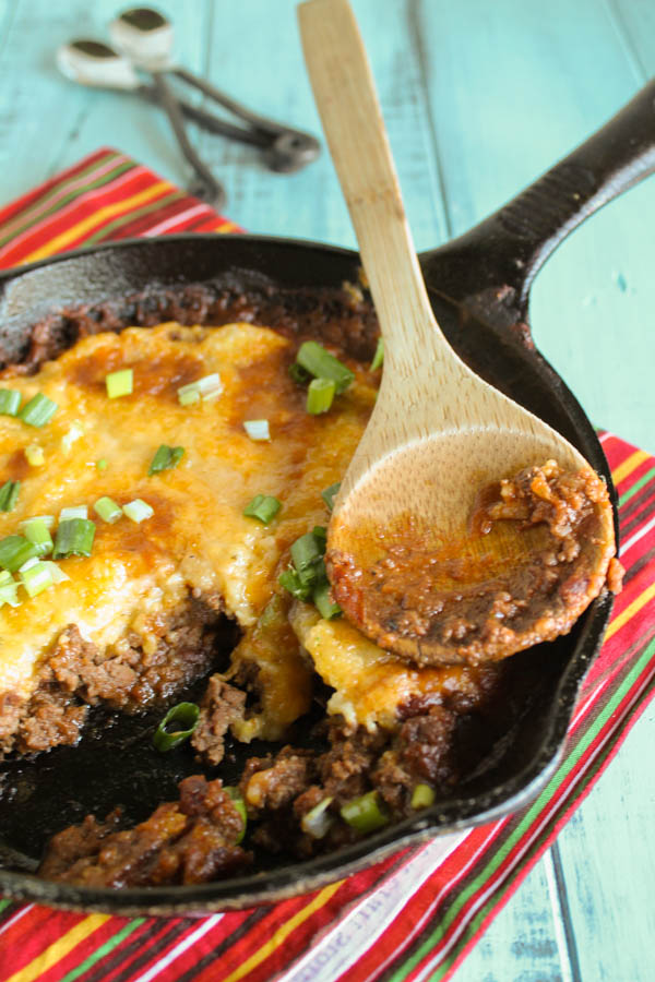 cast iron skillet filled with baked ground beef with a chewy tamale topping and a wooden spoon