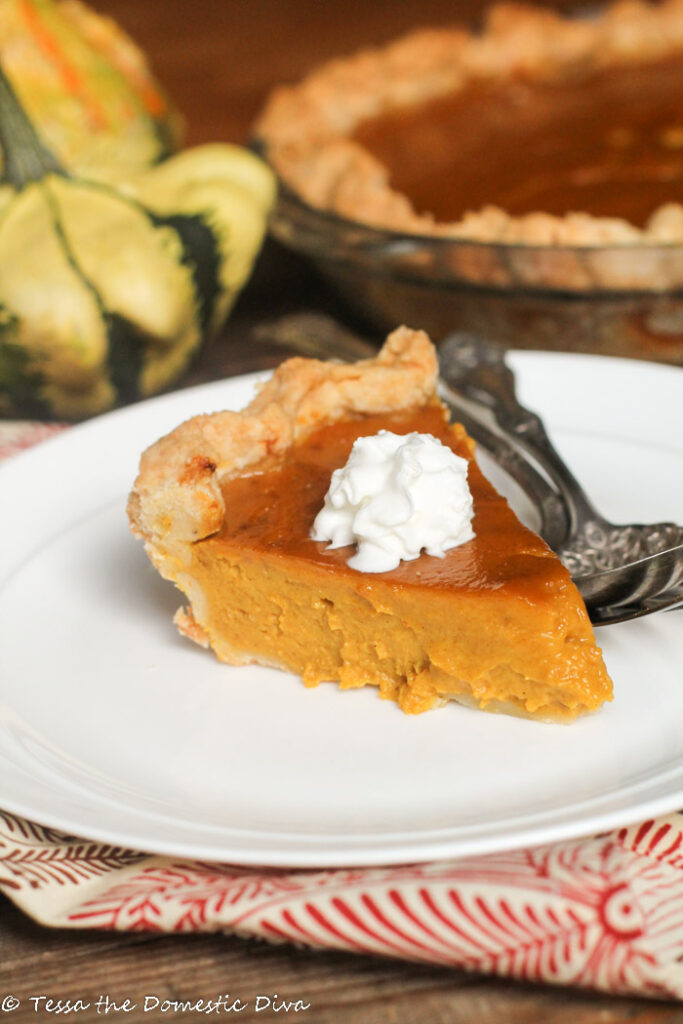 a slice of pumpkin pie with a dollop of whipped cream on a white plate with fall squash and a full pie in the background