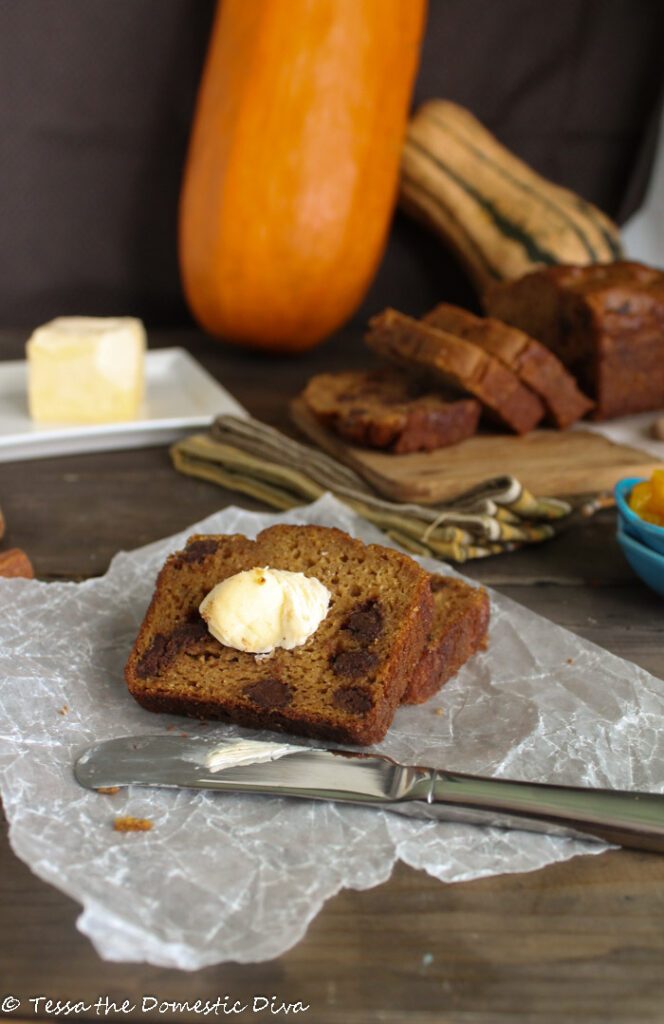 sliced pumpkin bread with a slather of butter o a parcemnt paper and dark wood surface with fresh pumpkin