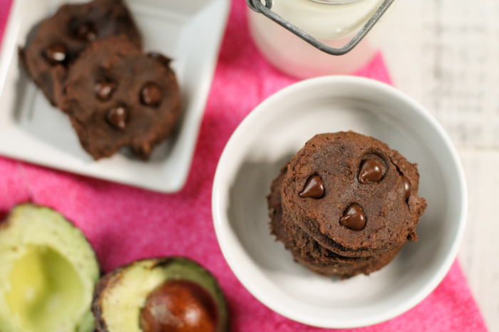 birdseye view of a white bowl filled with a stack of chococlate chip studded chococlate cookies, a vintage glass of milk, and an avocado half atop a pink linen