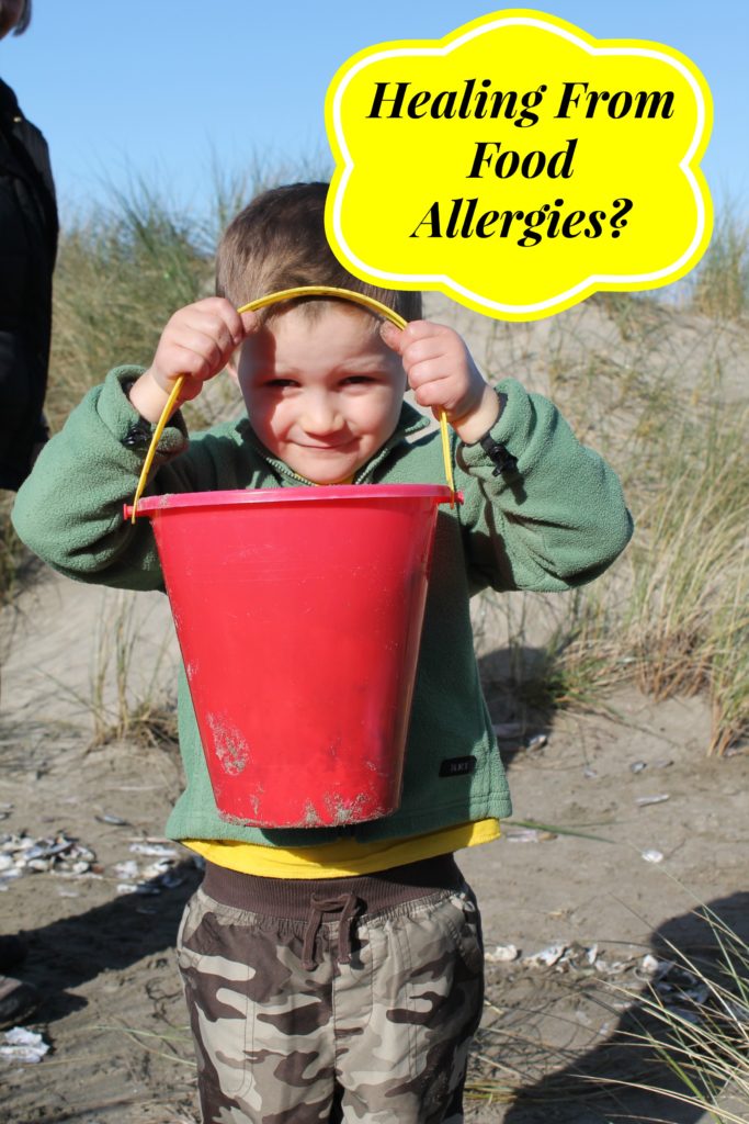 a 3 year old boy on a sunny beach with a red plastic bucket help up to his face