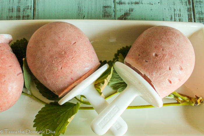 three strawberry cream popsicles from overhead arranged on a white plate with strawberry leaf garnish.