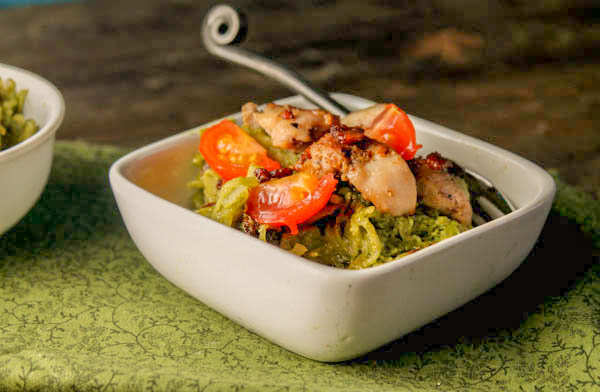 a square white bowl filled with pesto coated spaghetti squash noodles with chicken thighs and bacon