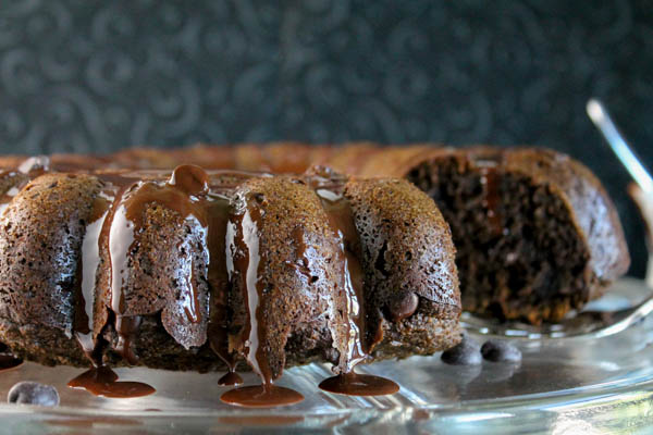 a sliced chocolate bundt cake with melted chocolate drizzled over the top on a clear glass cake plate with a dark background