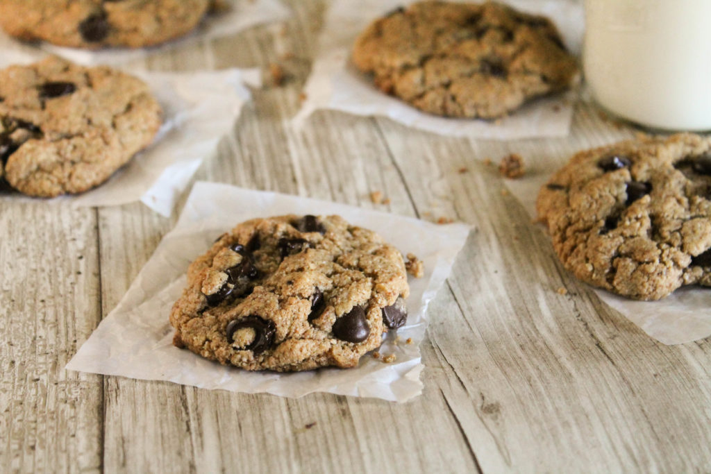 horizontal image of flourless paleo chococlate chip cookies arranged on a wooden surface on individual squares of parchment paper and a glass of milk