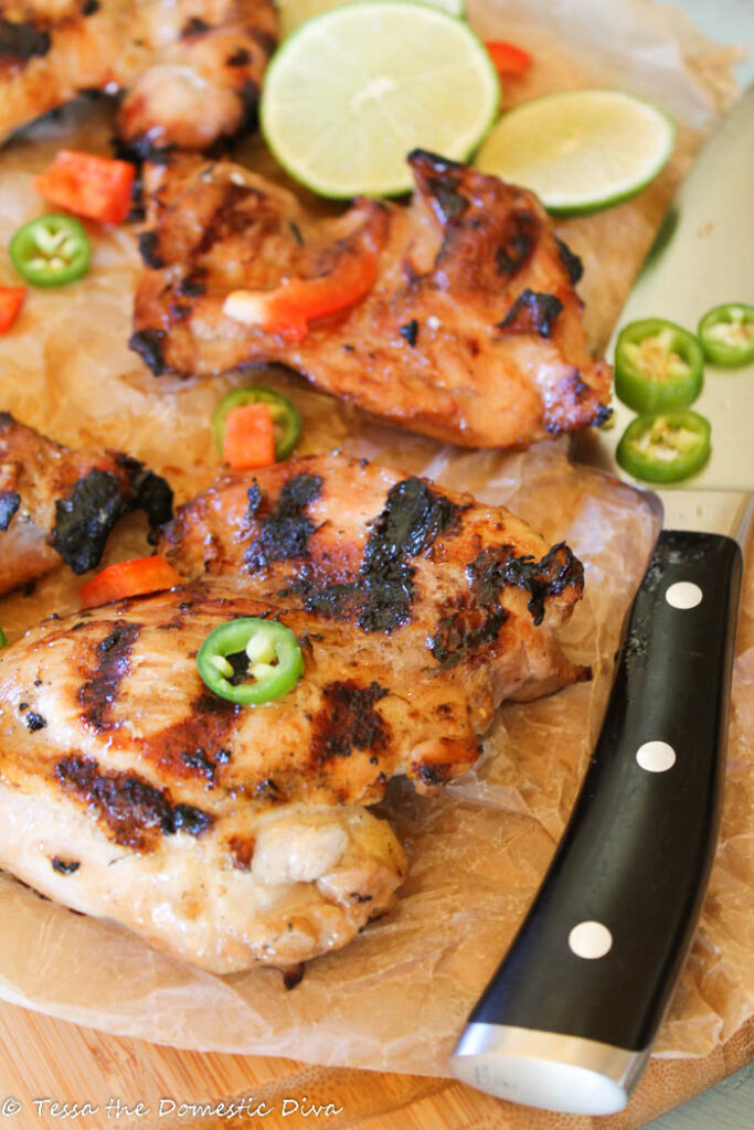 several grilled chicken thighs on top of a wooden cutting boardwith a black handled chef's knife and jalepeños