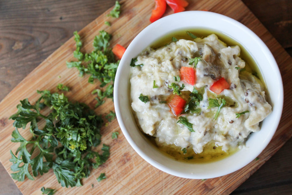 a white bowl filled with smoky eggplant dip topped with diced red peepers, parsley, and olive oil on a wooden cutting board