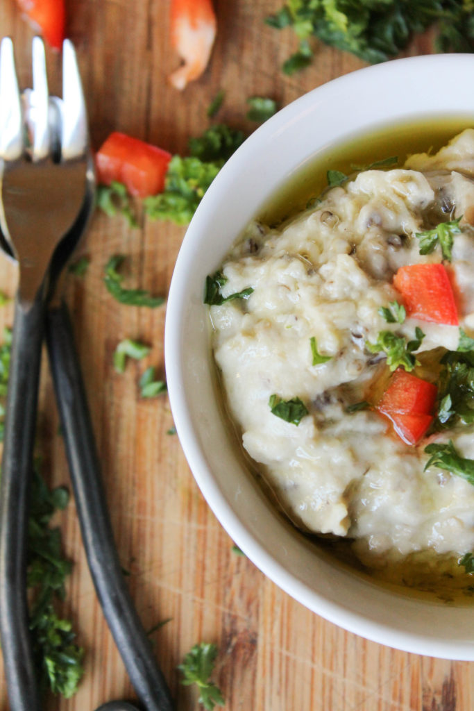 eggplant dip in a white bowl topped with diced red bell pepper, parsley, and olive oil from overhead.