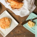 a fluffy biscuit on a white plate from overhead on a dark wooden surface with a turquoise butter dish