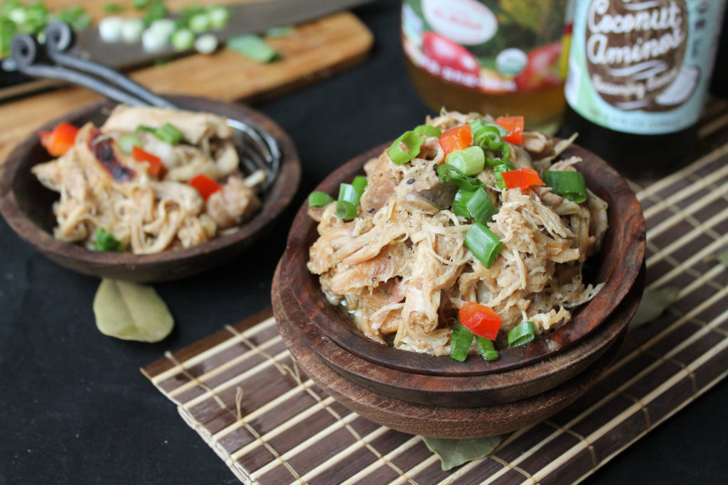 horizontal image of two dark wooden bowl filled with tender shredded adobo chicken on a bamboo mat with coconut aminos and vinegar bottle in background