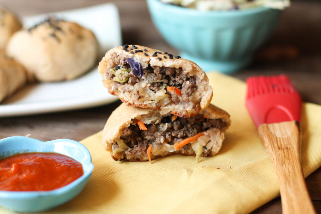 horizontal image of halved gluten free paleo baked bao buns filled with a savory hamburger filling on a pale yellow fabric with a turquoise dipping bowl of sriracha