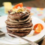 a stack of 6 wholegrain pancakes topped with a pat of butter, sliced almonds, and orange wedges on a white plate atop a dark wood surface