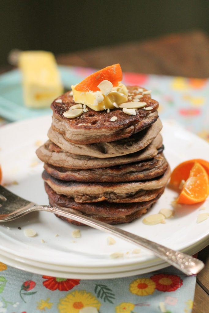 a large stack of brown teff pancakes with fresh oranges and sliced almonds on a white plate