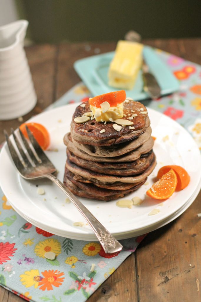 vertical image of a stack of dark brown pancakes topped with a pat of butter and orange wedges on a white plate with a blue, yellow, and orange floral fabric on dark wooden surface