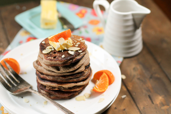a big stack of teff pancakes on a white plate with oranges, almonds, and a cube of butter