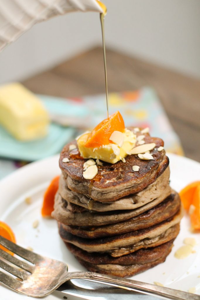 stack of dark brown pancakes topped with fresh butter and orange wedge and a drizzle of syrup atop a white plate