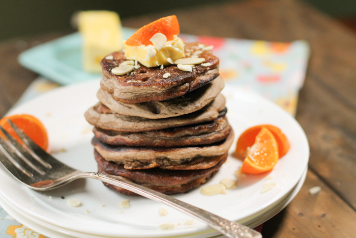 a stack of 6 wholegrain pancakes topped with a pat of butter, sliced almonds, and orange wedges on a white plate atop a dark wood surface