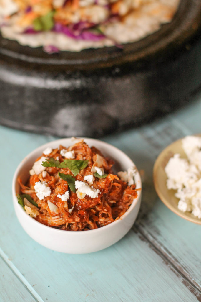 a white bowl with shreeded chicekn thighs in a tomaot chipotle sauce with crumbled feta. cilantro and cast iron pan in background