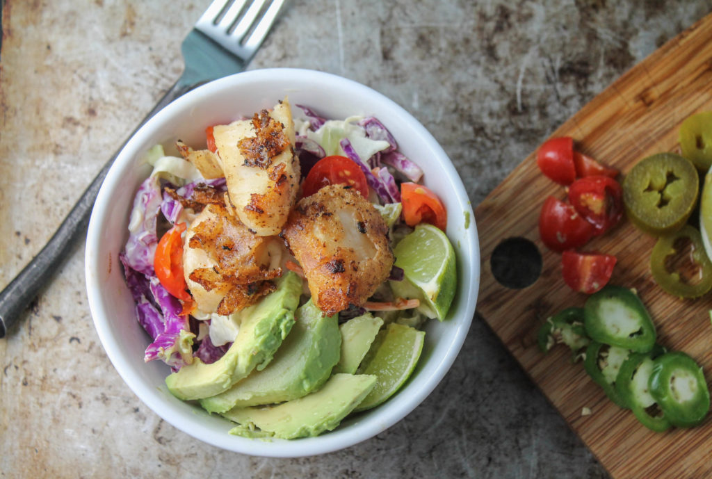 birdseye view of a white bowl filled with green and purple cabbage slaw topped with crispy breaded fish, avocados. and fresh lime