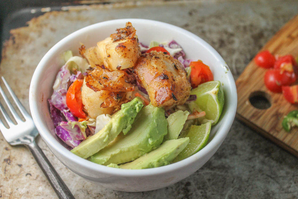 a white bowl of slaw and crispy fish tacos atop an aged cookie sheet with fresh avocado and jalapenos