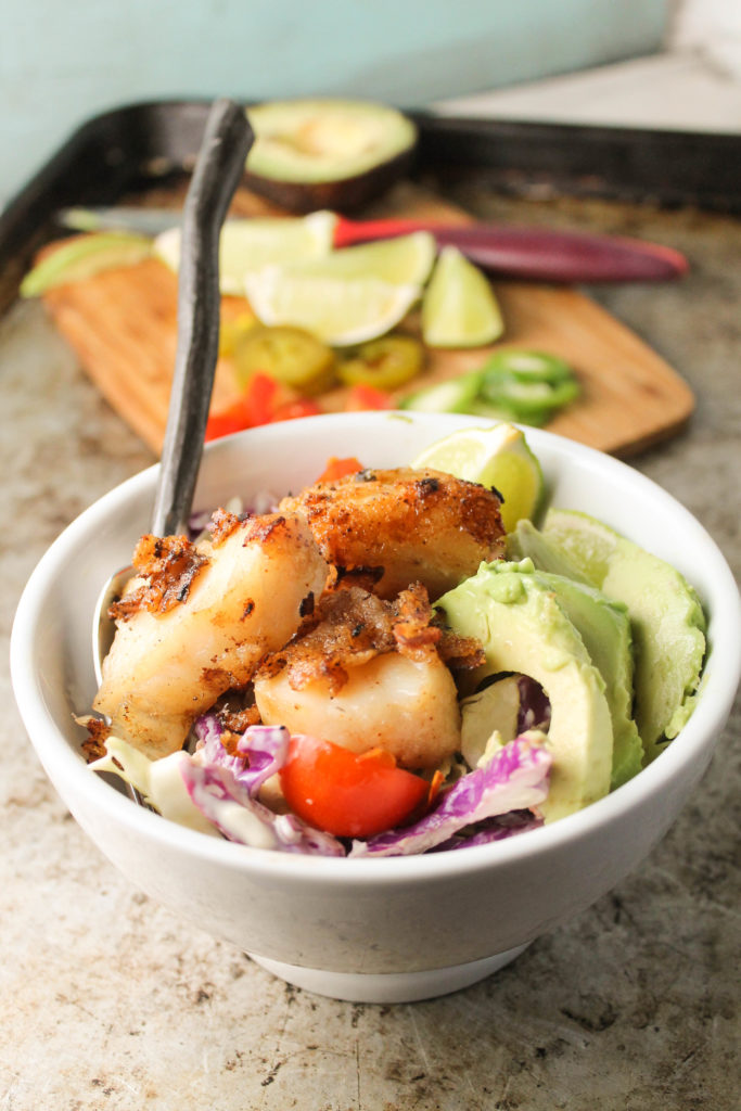 looking on to a white bowl filled with colorful slaw topped with crispy fish pieces, avocado, and cutting board in background