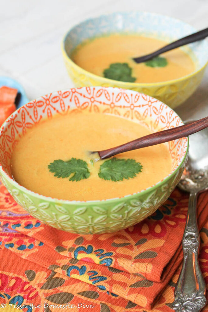 two festive bowls filled with a Thai inspired creamy carrot soup with cilantro, chilies, and olive wood spoons