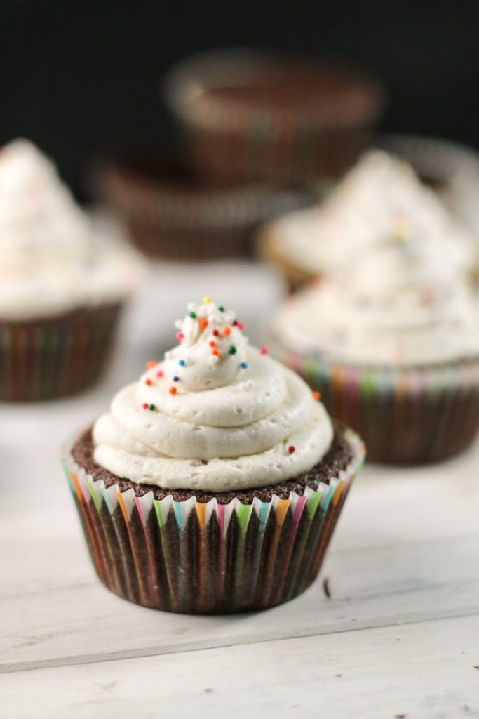 vertical image of a chocolate cupcake in a colorful paper wrapper with a swirl of white buttercream frosting and sp