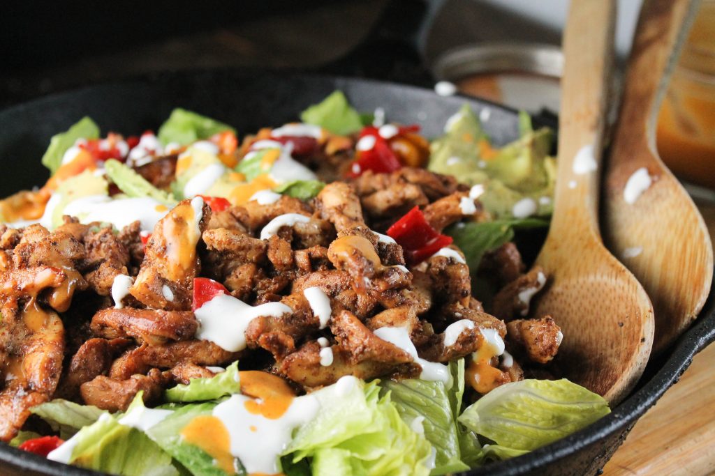 frontal view of savory chciem fajitas in a cast iron pan with wooden serving spoons, sauteed onions, peppers, and sliced avocado