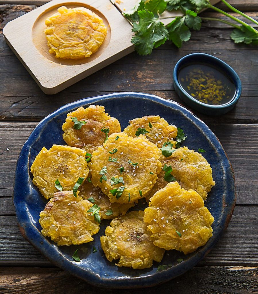 dark blue pottery plate topped with fried green plantains and sprinkle f salt on a dark wooden surface