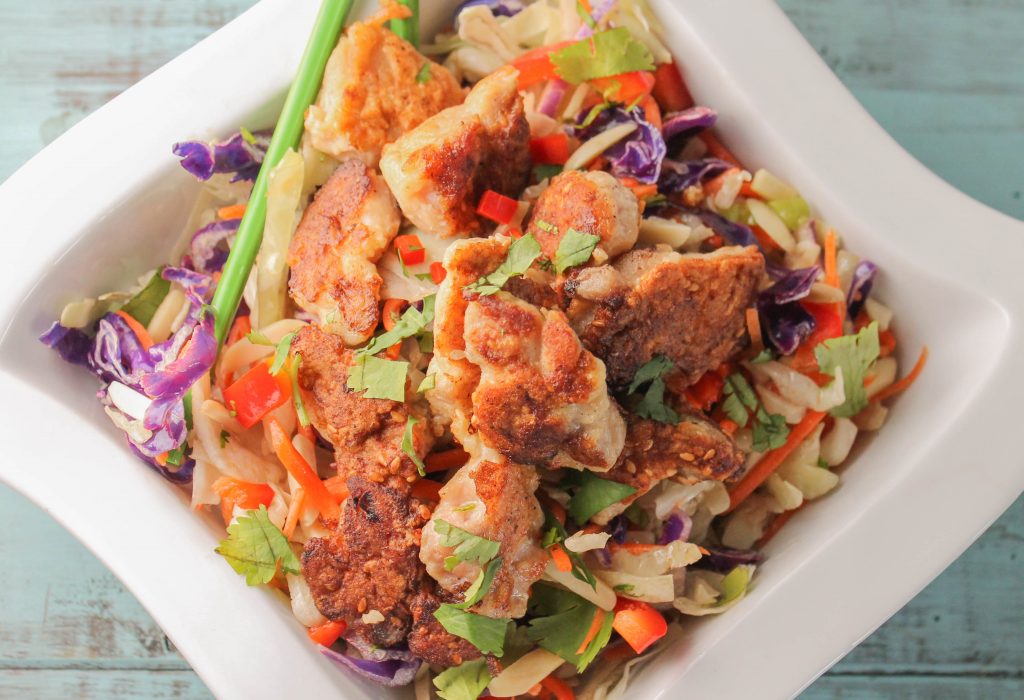 horizontal image of crispy sesame coated chicken pieces atop a bed of cabbage slaw and red peppers with a garnish of cilantro