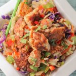 square white bowl from overhead on light blue wooden board filled with cruncy fried chicken peices with a a bright cabbage slaw and green chopsticks