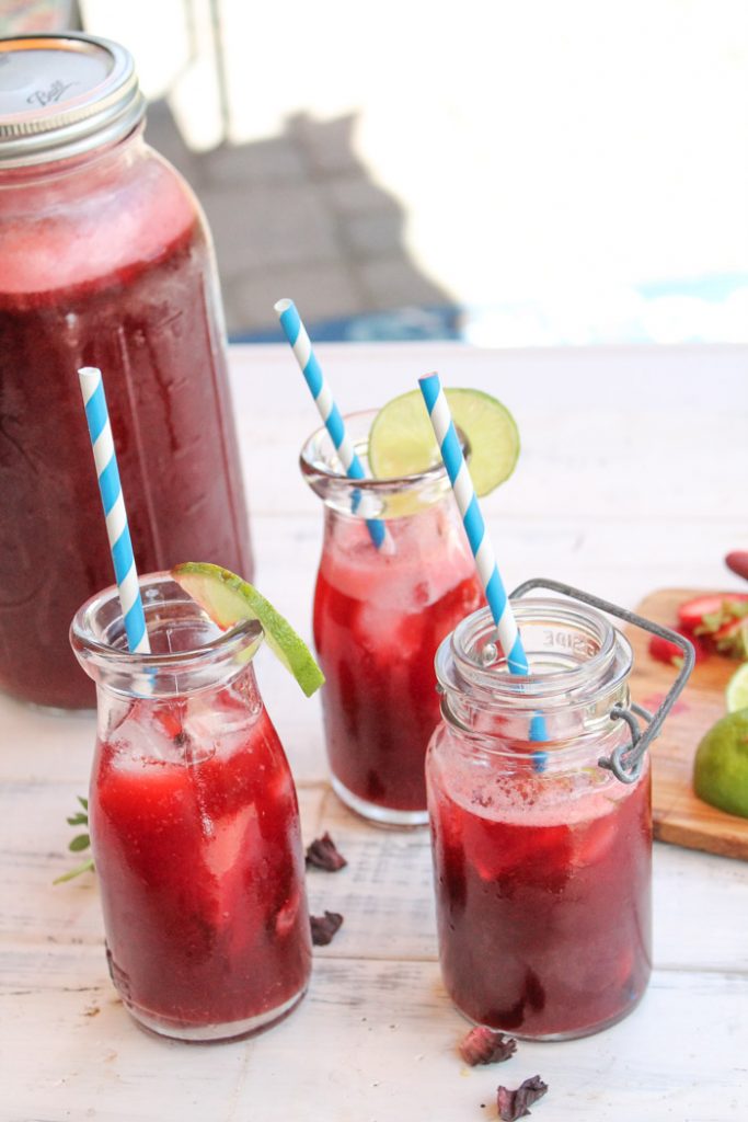 vertical image of 3 clear vintage glass milk glasses with blue and white striped paper straws filled with a vivid red tea with sliced strawberries, lime slices, and hibiscus flowers