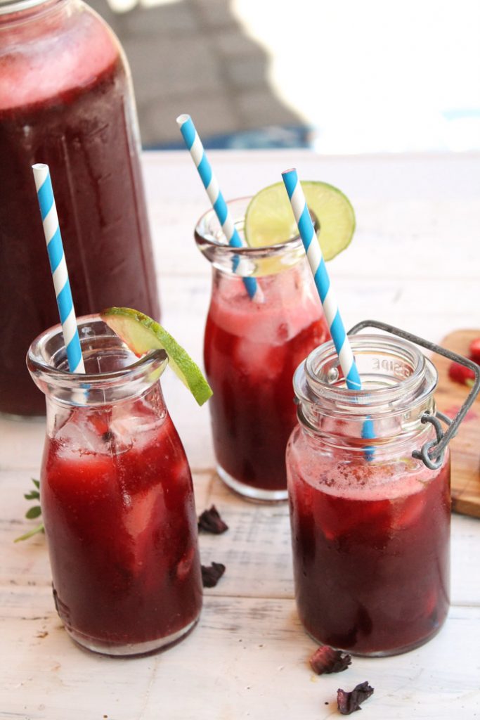 3 glasses of deep red hibiscus lime strawberry tea on a white board with large mason jar pitcher in background