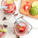 overhead shot of three clear glass cups with blue and white striped paper straw filled with vibrant red hibiscus tea, lime wedges, and sliced strawberries