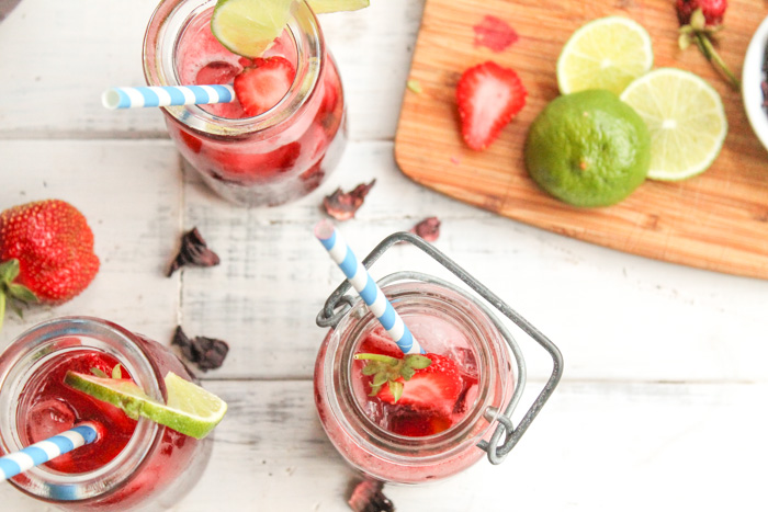 overhead shot of three clear glass cups with blue and white striped paper straw filled with vibrant red hibiscus tea, lime wedges, and sliced strawberries