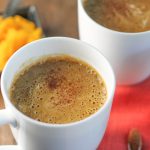 a white mug filled with a orange hued frothy beverage topped with a sprinkle of cinnamon on a fall coloered linen and fresh pumpkin puree in the background on a dark wood surface