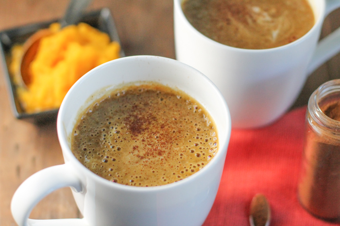 a white mug filled with a orange hued frothy beverage topped with a sprinkle of cinnamon on a fall coloered linen and fresh pumpkin puree in the background on a dark wood surface