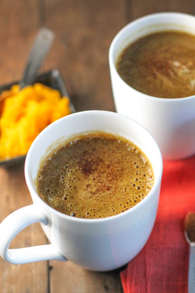 two white mugs filled with an orange hued coffee drink with a dish of fresh pumpkin puree and a fall colored linen
