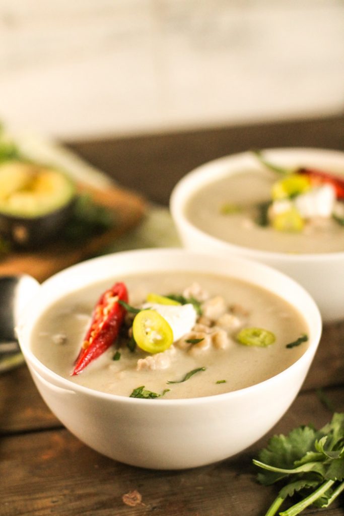 two white bowls on a dark wooden surface filled with a creamy white soup garnished with avocado, sour cream, and sliced jalapenos