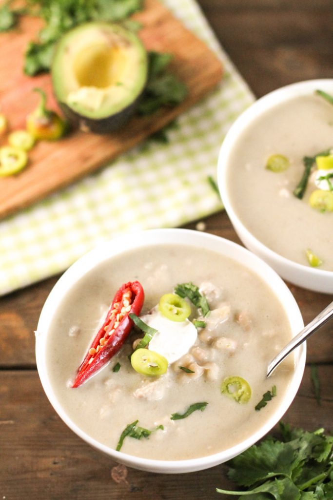 two white bowls from overhead filled with a creamy white soup and garnished with fresh cilantro, red pepper, and sour cream on a dark wood surface