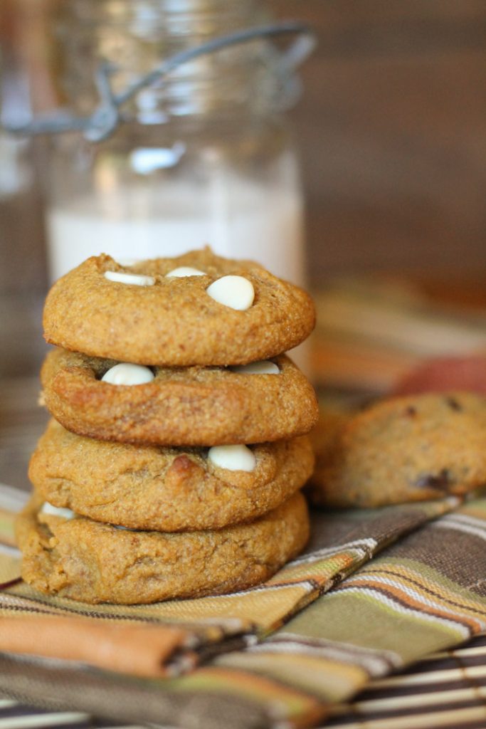 a stack of 4 thick and chewy orange hued pumpkin cookies studded with white chcocolate chips on a fall colored napkin with a vintage milk glass in the background