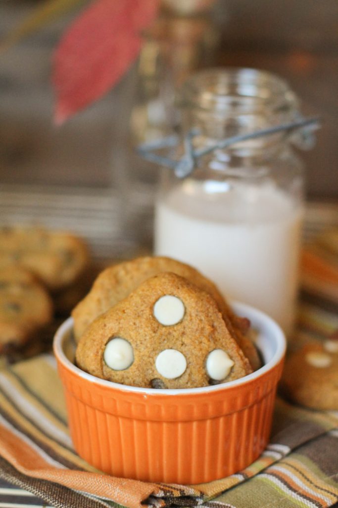 a few white chocolate studded pumpkin cookies standing in pumpkin orange ramekin with a glass of milk in background