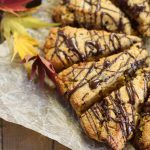 horizontal image of chocoalte drizzled pumpkin scones from overhead atop a crumpled parchment paper and fall leaves scattered about