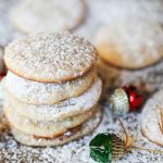 horizontal image of 5 stacked cheesecake cookies sprinkled with powdered auagr on a cookie sheet with a garlnad of red and green ornaments scattered about