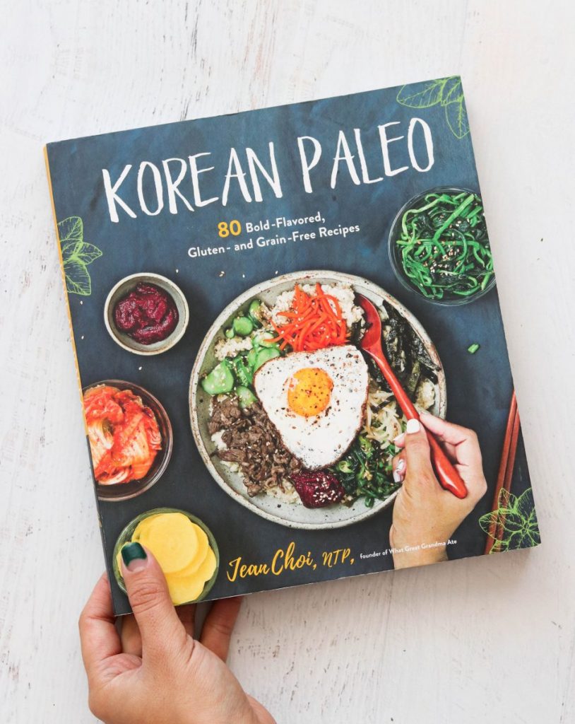 vertical image of korean paleo cookbook from overhead on a light surface with a hand holding the book in the left hand corner