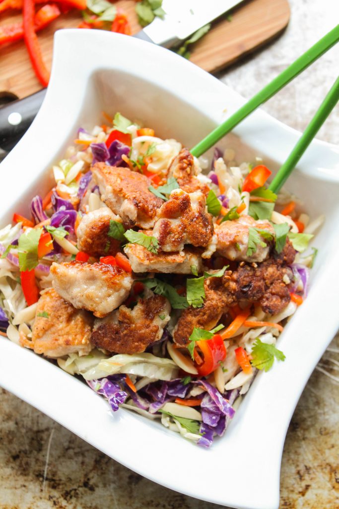 vertical image of a sqaure white bowl filled with cabbaje slaw, fresh red pper, and crispy sesame coated chicken bites with green chopsticks atop a worn steel cookie sheet